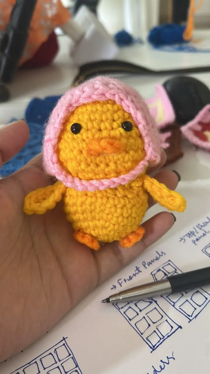 A yellow amigurumi chick in a pink bonnet, held in a hand